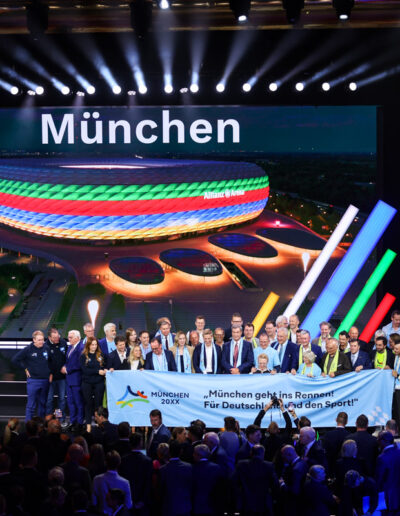 Gruppenfoto zur Olympiabewerbung München 20XX: Vertreter aus Sport und Politik präsentieren auf der Bühne ein Banner mit dem Motto ‚München geht ins Rennen!‘. Im Hintergrund ist die farbenprächtig beleuchtete Allianz Arena zu sehen.