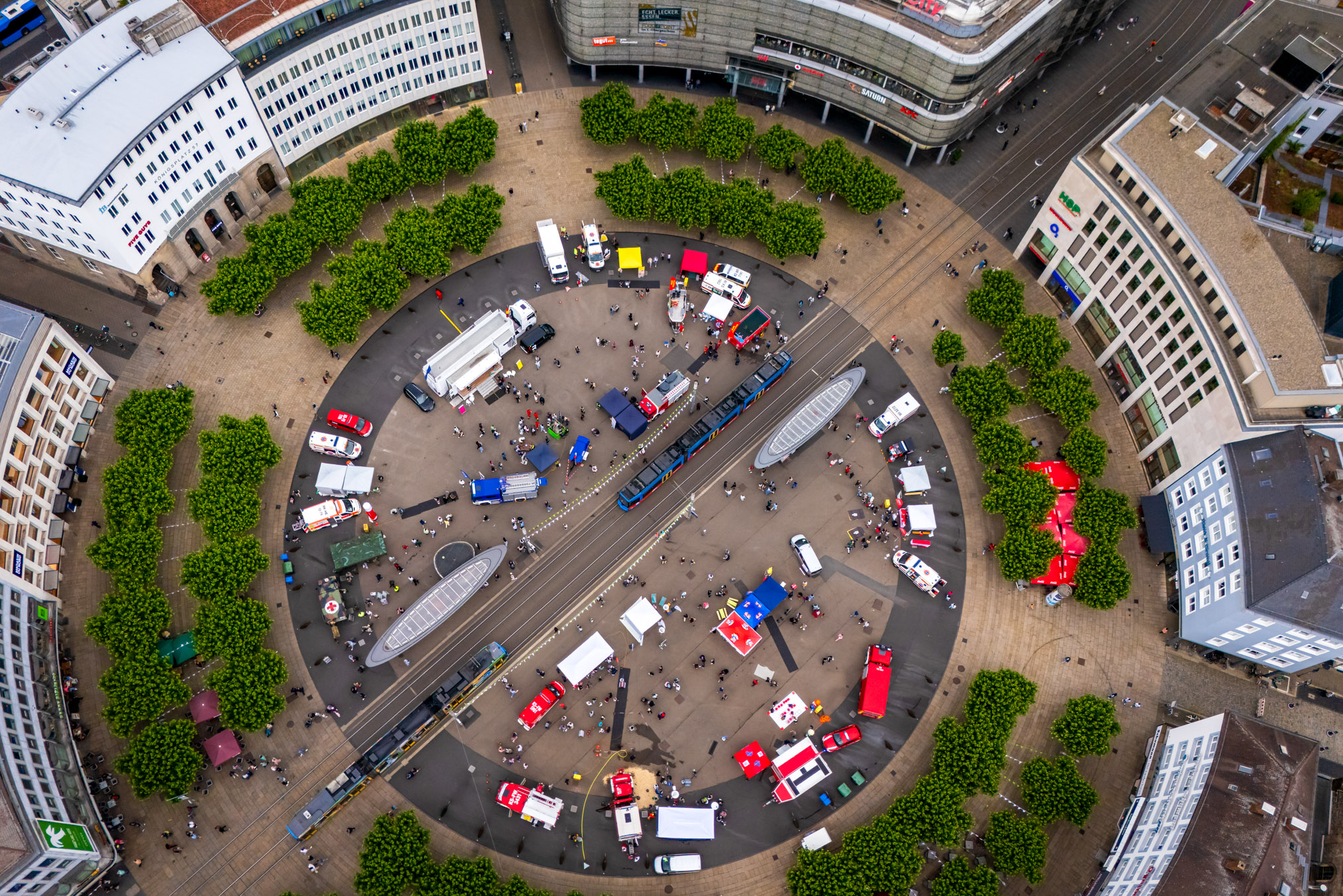 Drohnenaufnahme einer Veranstaltung zum Bevölkerungsschutz auf einem großen, kreisförmigen Stadtplatz mit Feuerwehr- und Rettungsfahrzeugen, Informationsständen und zahlreichen Besuchern. Straßenbahnen durchqueren den Platz.