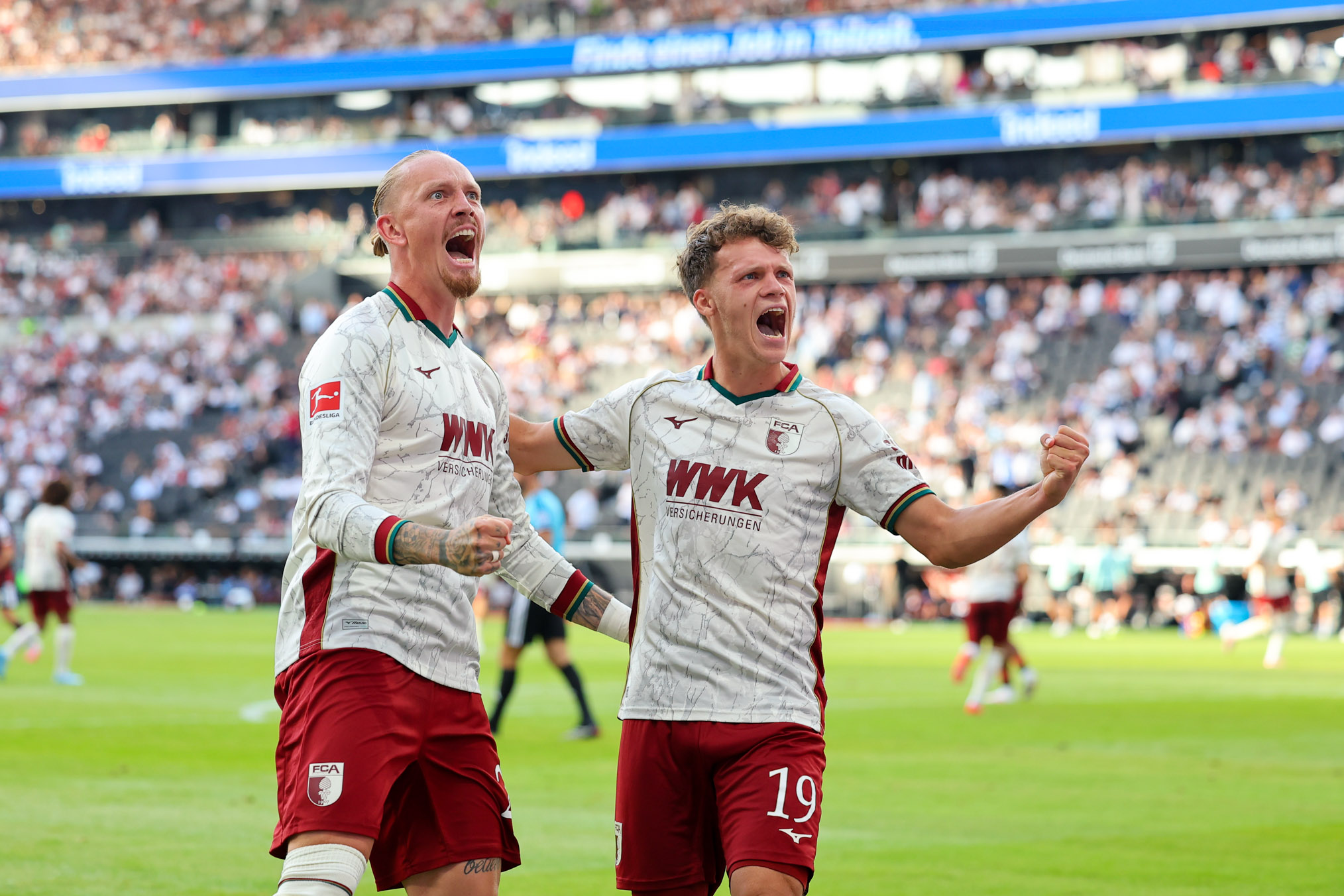 Zwei Fußballspieler des FC Augsburg jubeln gemeinsam mit geballten Fäusten und emotionalen Gesichtsausdrücken während eines Bundesligaspiels vor gefüllten Stadionrängen.